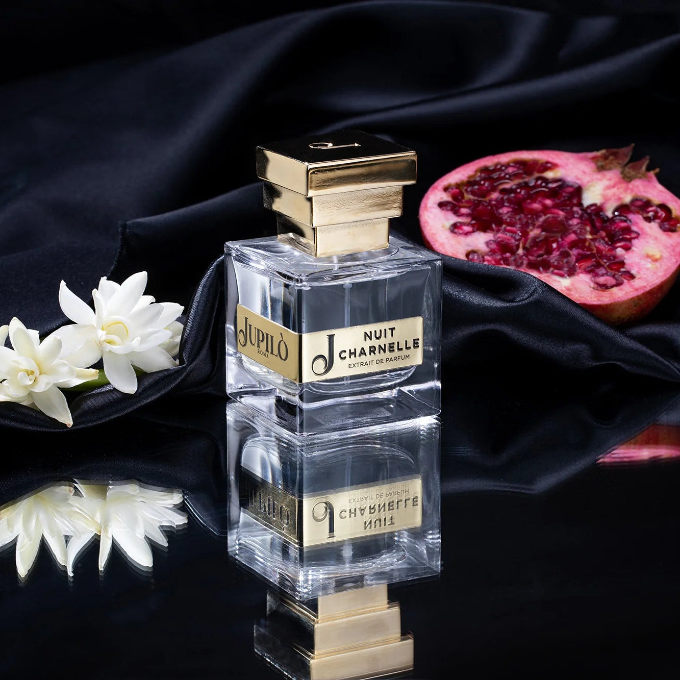 Flacone di profumo Nuit Charnelle su tessuto nero vicino a fiori bianchi e melograno