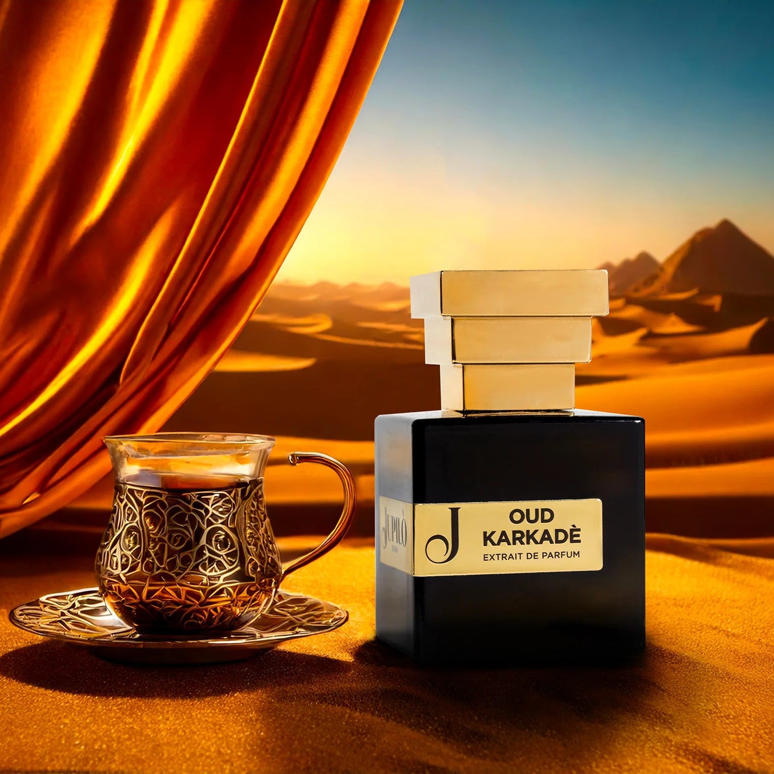 Flacone di profumo Oud Karkadè accanto a bicchiere decorato, sfondo deserto dorato.