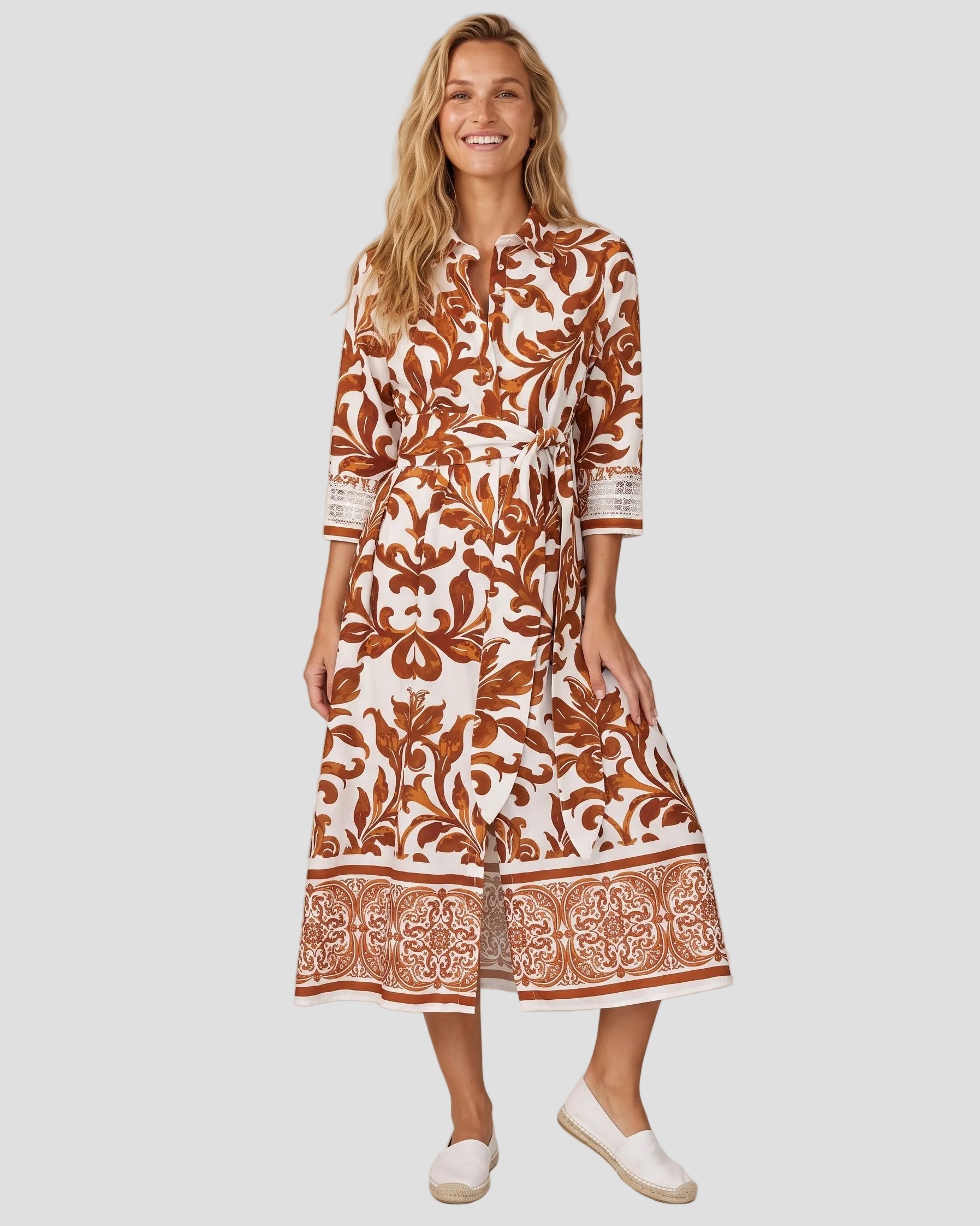  Modella sorridente indossa l'abito chemisier midi Milva MI in cotone bianco e marrone terra bruciata con stampa maiolica e scarpe espadrilles bianche.