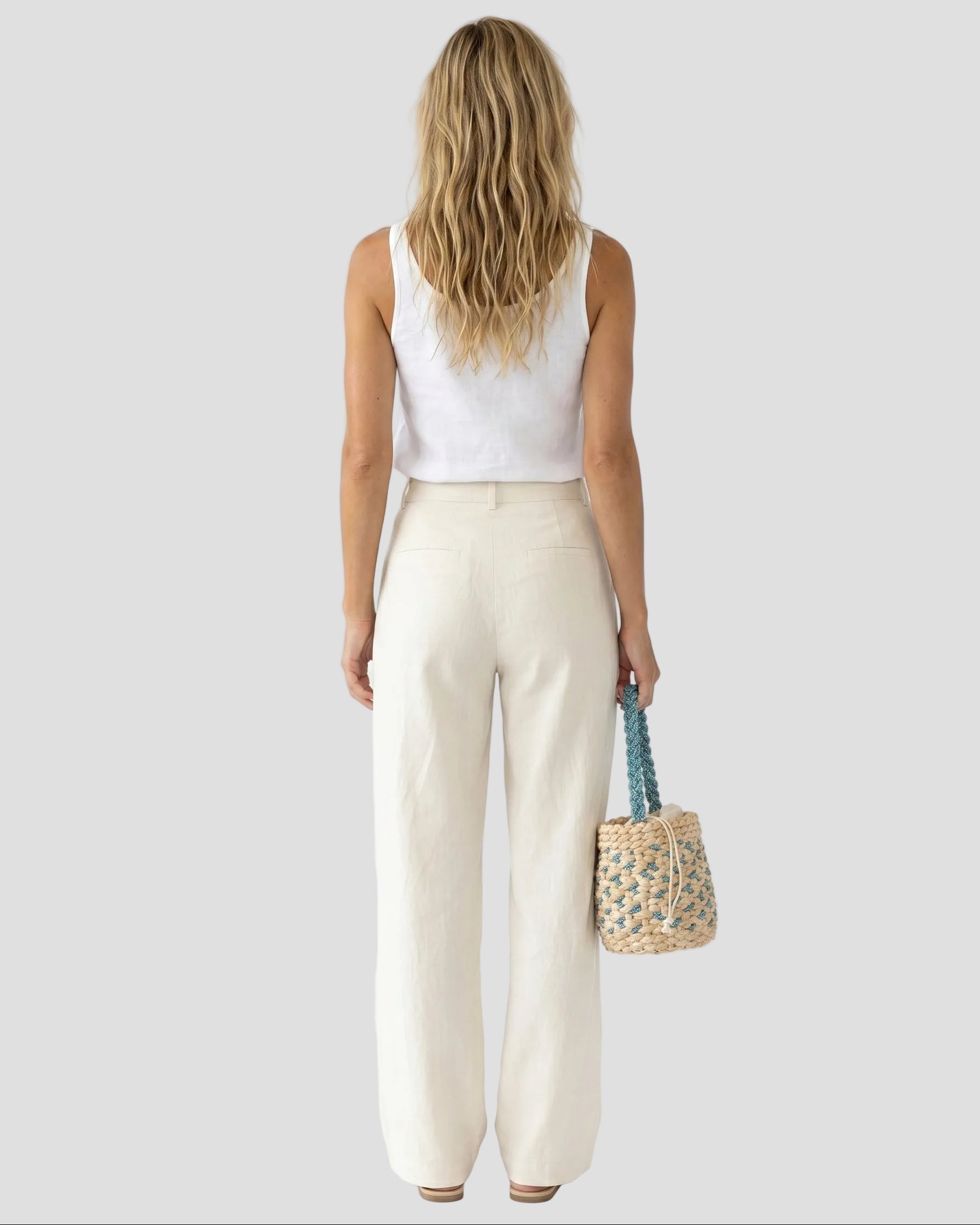 Modella di spalle con capelli biondi mossi, top bianco e pantaloni in lino panna, che tiene in mano la borsa a secchiello in rafia azzurra Pho Firenze.