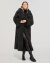 Donna con cappotto lungo oversize nero eco pelliccia, stivali alti neri, fondo neutro
