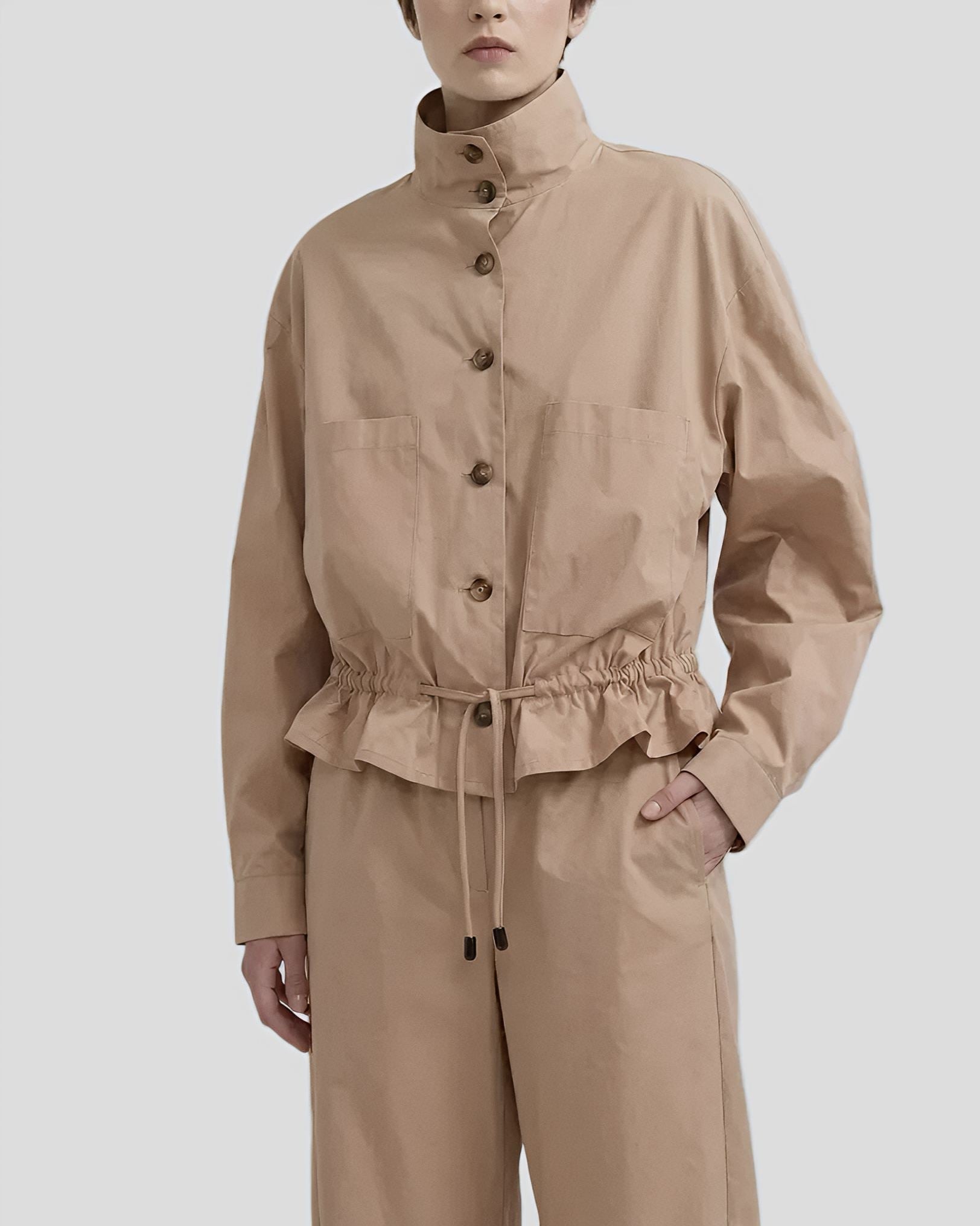 Dettaglio ravvicinato di un completo beige composto da giacca bomber con coulisse in vita e bottoni scuri, indossato da una modella con taglio corto."