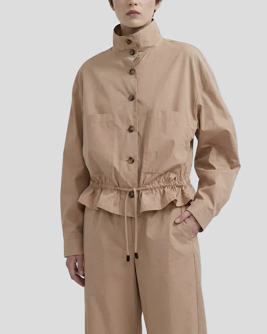 Dettaglio ravvicinato di un completo beige composto da giacca bomber con coulisse in vita e bottoni scuri, indossato da una modella con taglio corto."