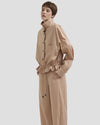 Donna che indossa una giacca stile sahariana beige oversize corta, abbinata a pantaloni larghi coordinati; il modello presenta un collo alto, tasche frontali e coulisse in vita