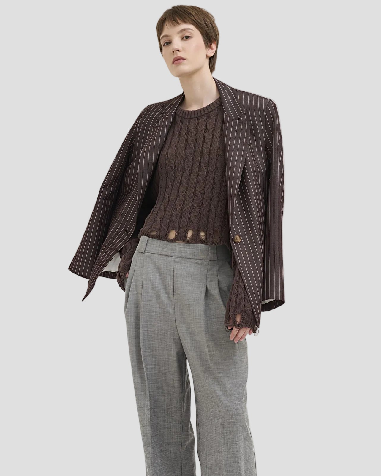 Modella con maglione traforato a trecce marrone, blazer a righe e pantaloni grigi larghi.